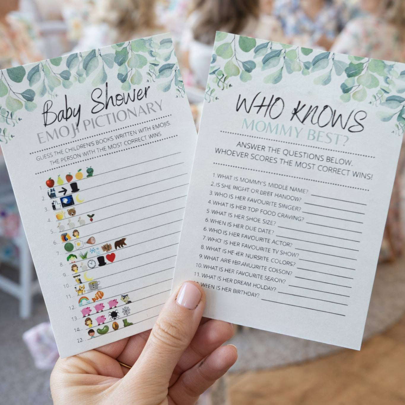 Two baby shower games held by a hand with a blurred background of people.