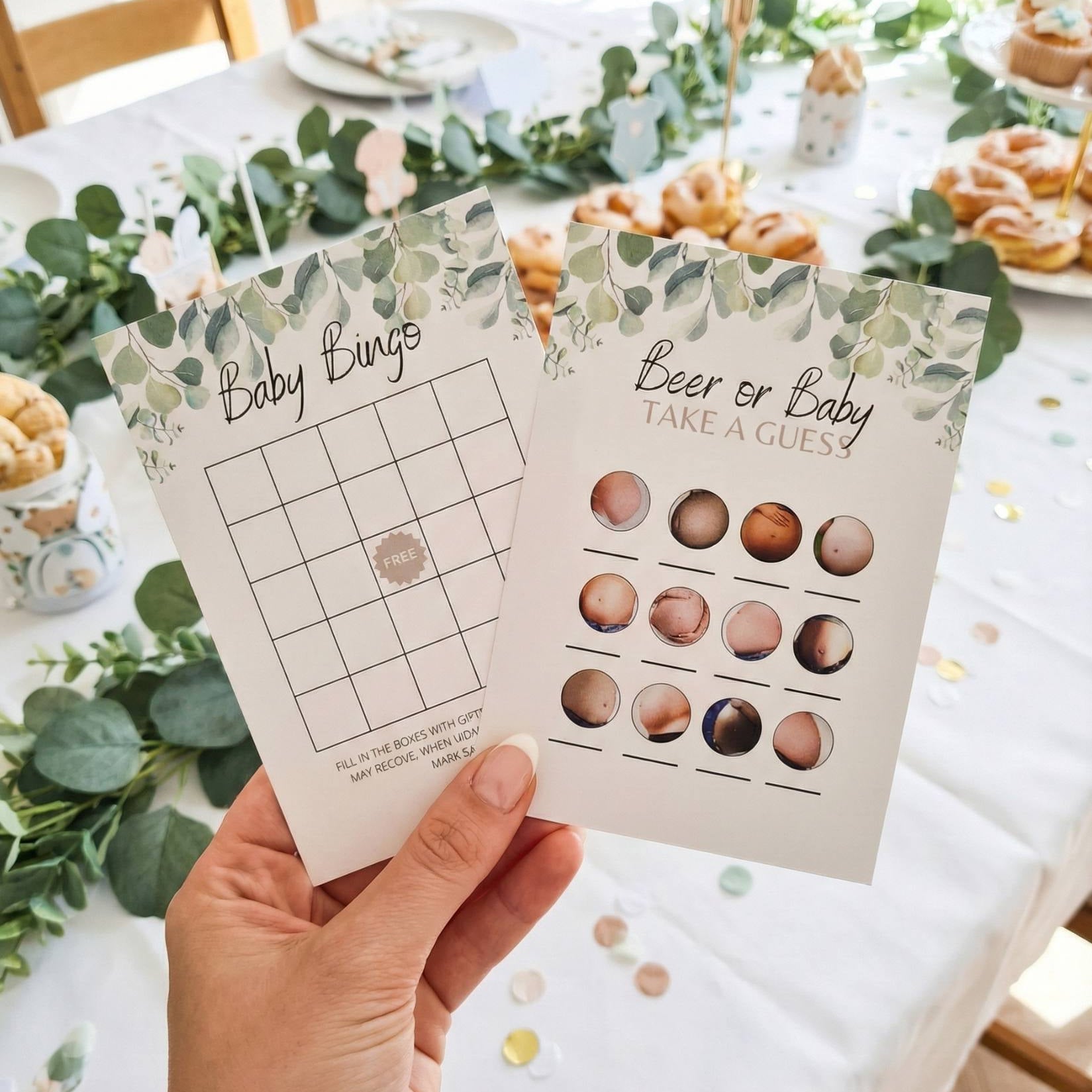 Hand holding two baby shower games on a decorated table with food and greenery.