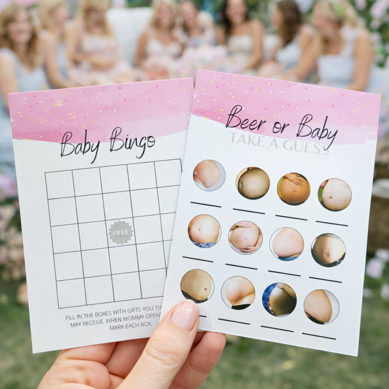 Two baby shower bingo cards held by a hand with a blurred outdoor party background.