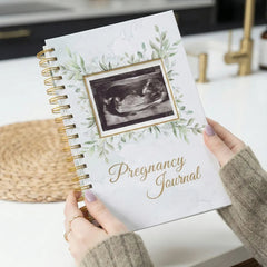 Pregnancy journal with ultrasound photo and greenery design held by a person in a kitchen.